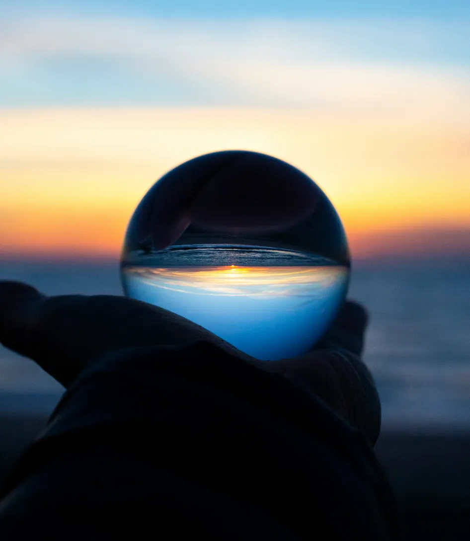 A hand holding a glass sphere that reflects an inverted seaside sunset.