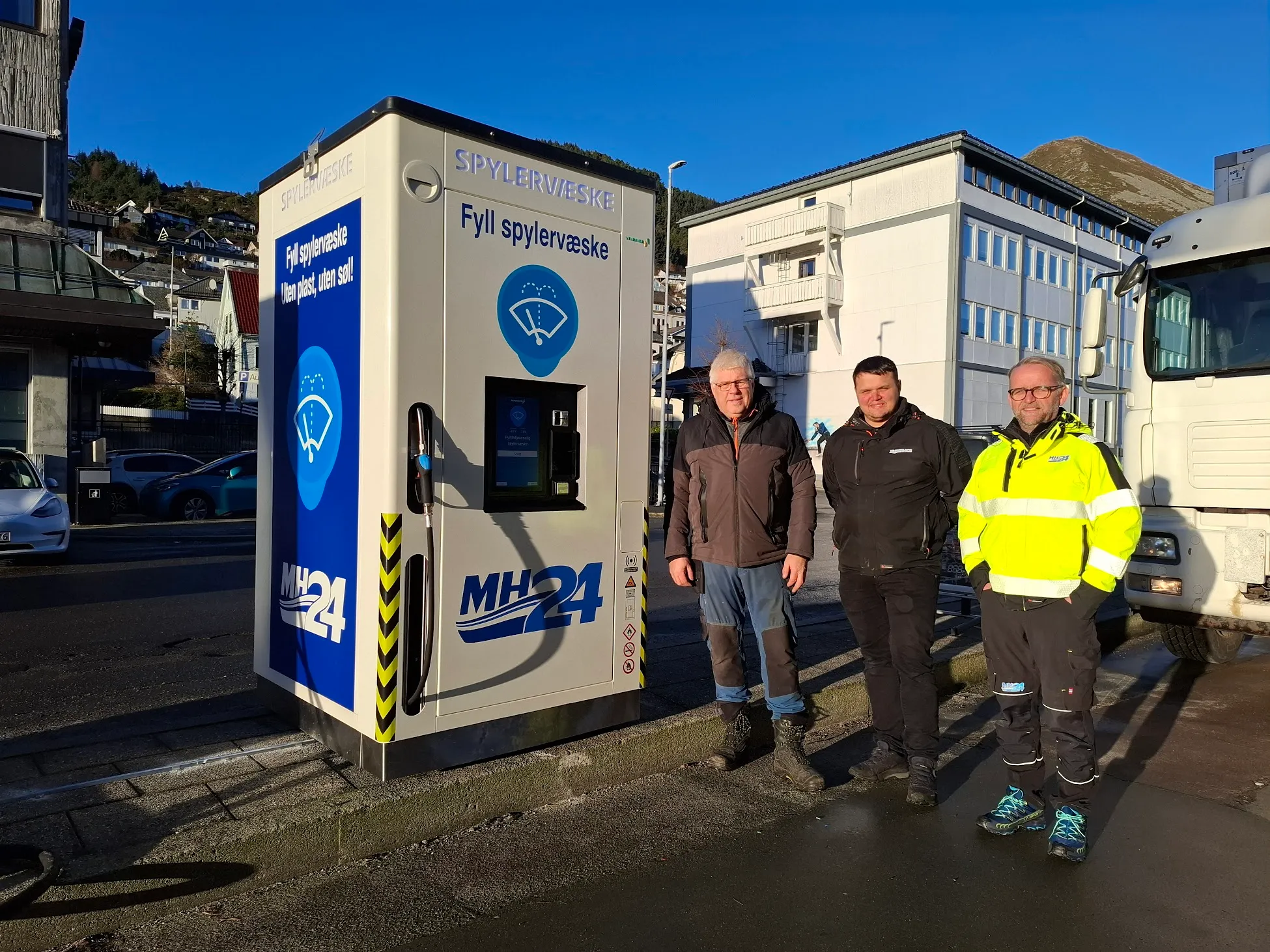 Three men standing beside an MH24-branded Vendanor Washer Fluid Dispenser on a street in Måløy.