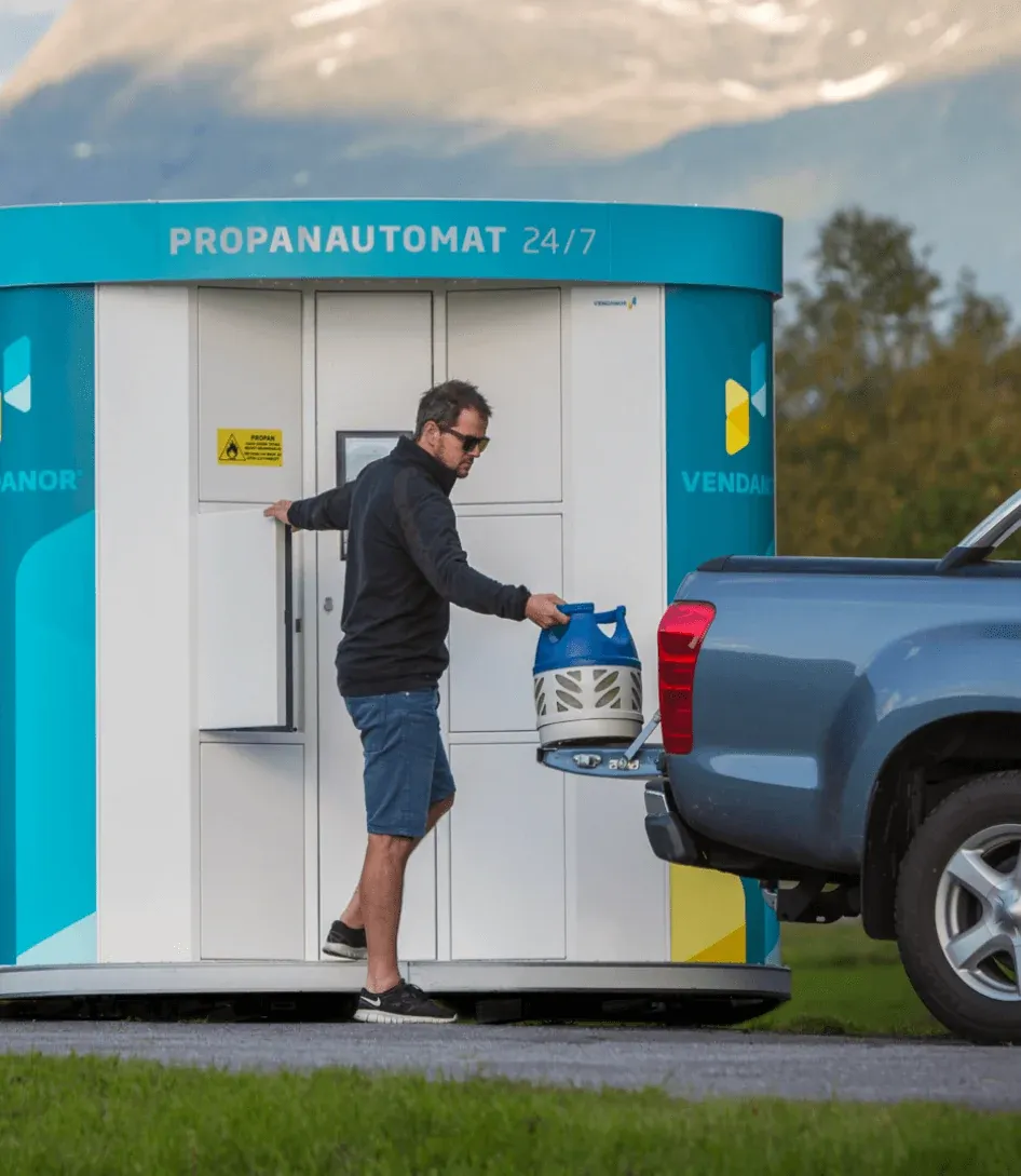 Man loading a propane cylinder from a Vendanor Propane Vending Machine into a pickup truck in a mountain landscape.
