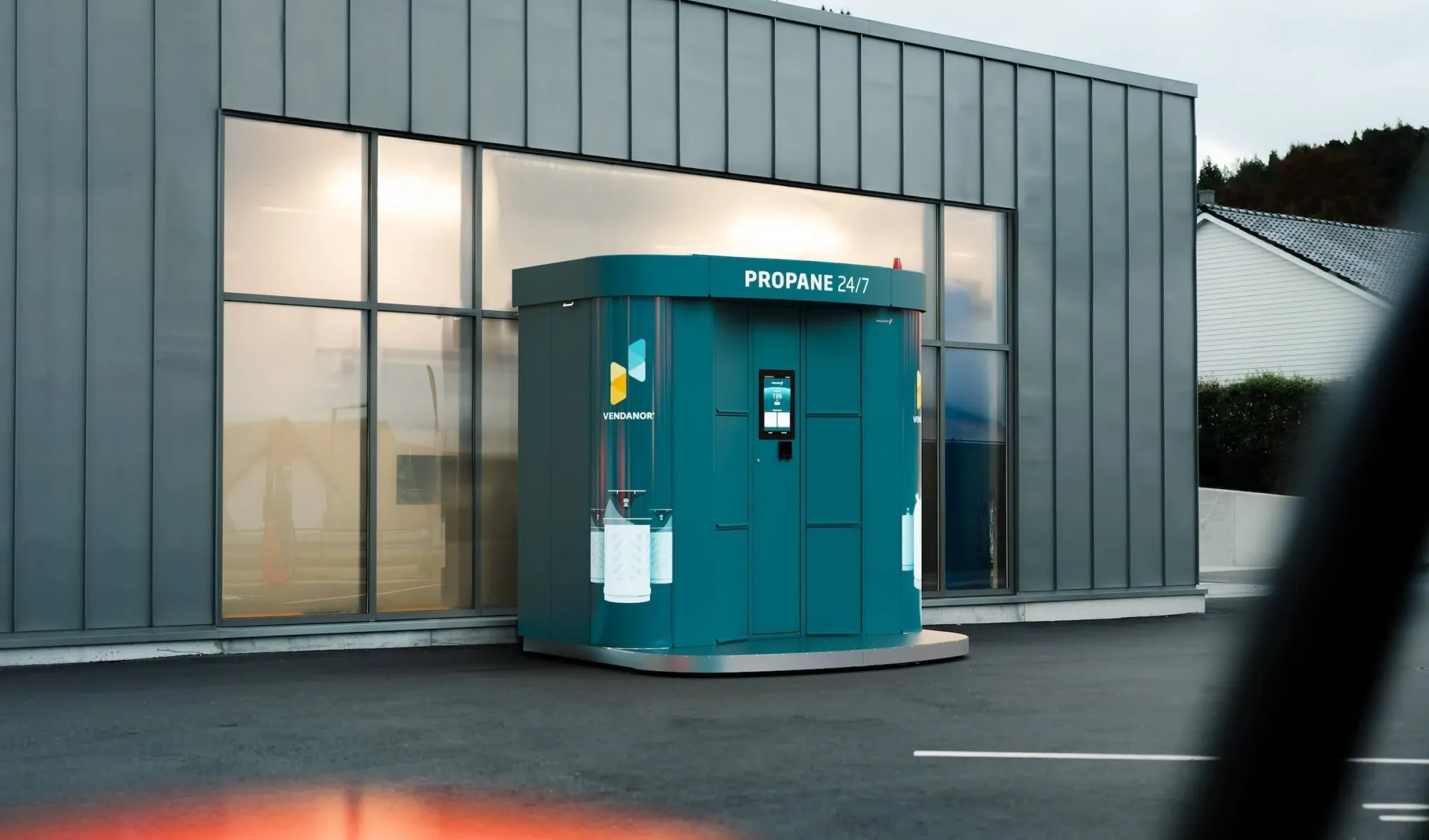 A teal Vendanor Propane Vending Machine labelled Propane 24/7 stands beside a grey industrial building, viewed past a car window.